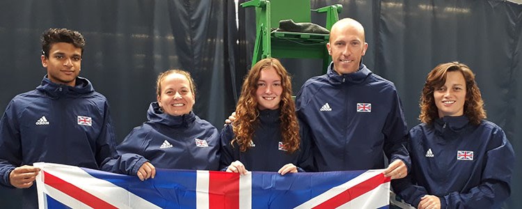 four tennis players and coach wearing GB blue jackets holding up  big GB flag