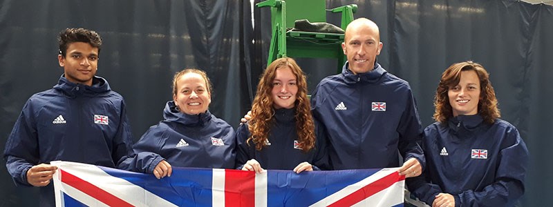 four tennis players and coach wearing GB blue jackets holding up  big GB flag