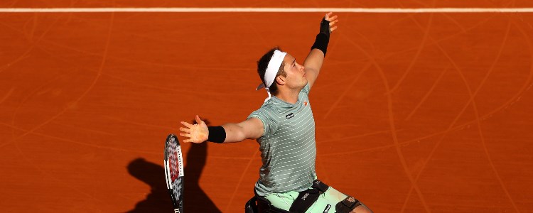 Alfie Hewett rejoicing with his hand in the air on a red tennis court