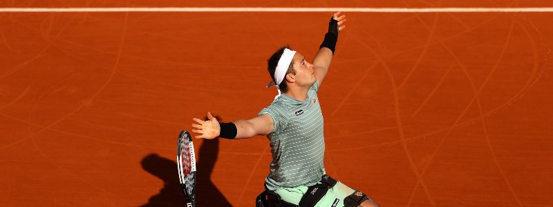 Alfie Hewett rejoicing with his hand in the air on a red tennis court