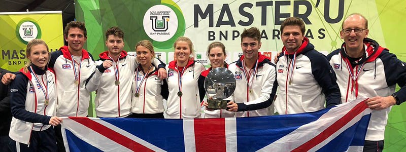 GB unviersity team standing in a line with medals and holing a trophy and holding big GB flag at the front