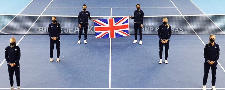 Six people standing a small distance apart on a tennis court with the back two holding a United Kingdom flag up