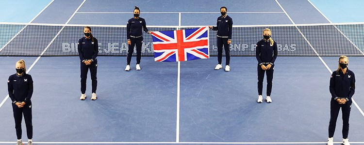 Six people standing a small distance apart on a tennis court with the back two holding a United Kingdom flag up