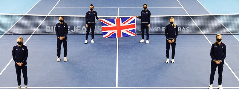 Six people standing a small distance apart on a tennis court with the back two holding a United Kingdom flag up