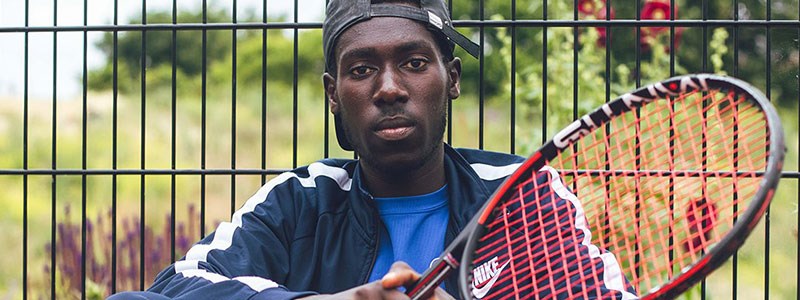 close up of Arum Akom sitting in front of a fence with tennis racket in hand