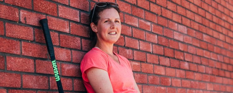 Elly Shearman leaning against a brick wall smiling with Wilson tennis racket next to her