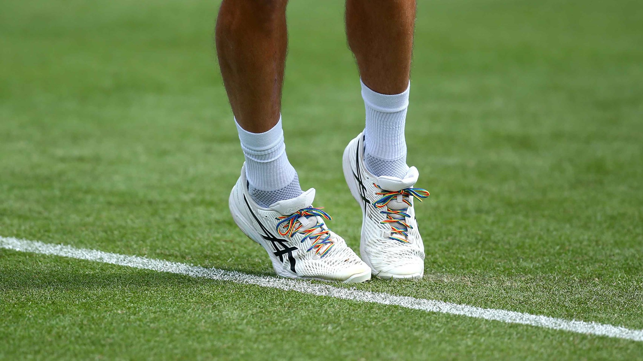 An image of a tennis player's legs, stood on a grass court, with rainbow coloured laces on their shoes