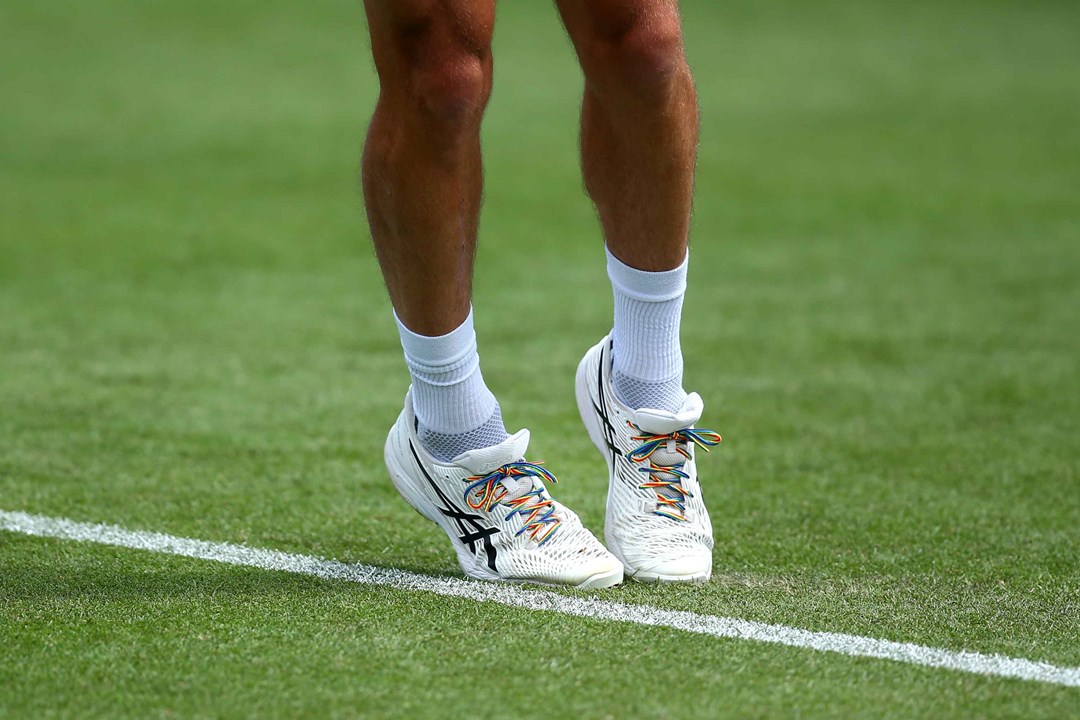 An image of a tennis player's legs, stood on a grass court, with rainbow coloured laces on their shoes