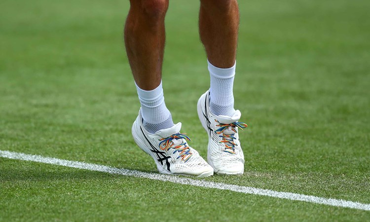 An image of a tennis player's legs, stood on a grass court, with rainbow coloured laces on their shoes