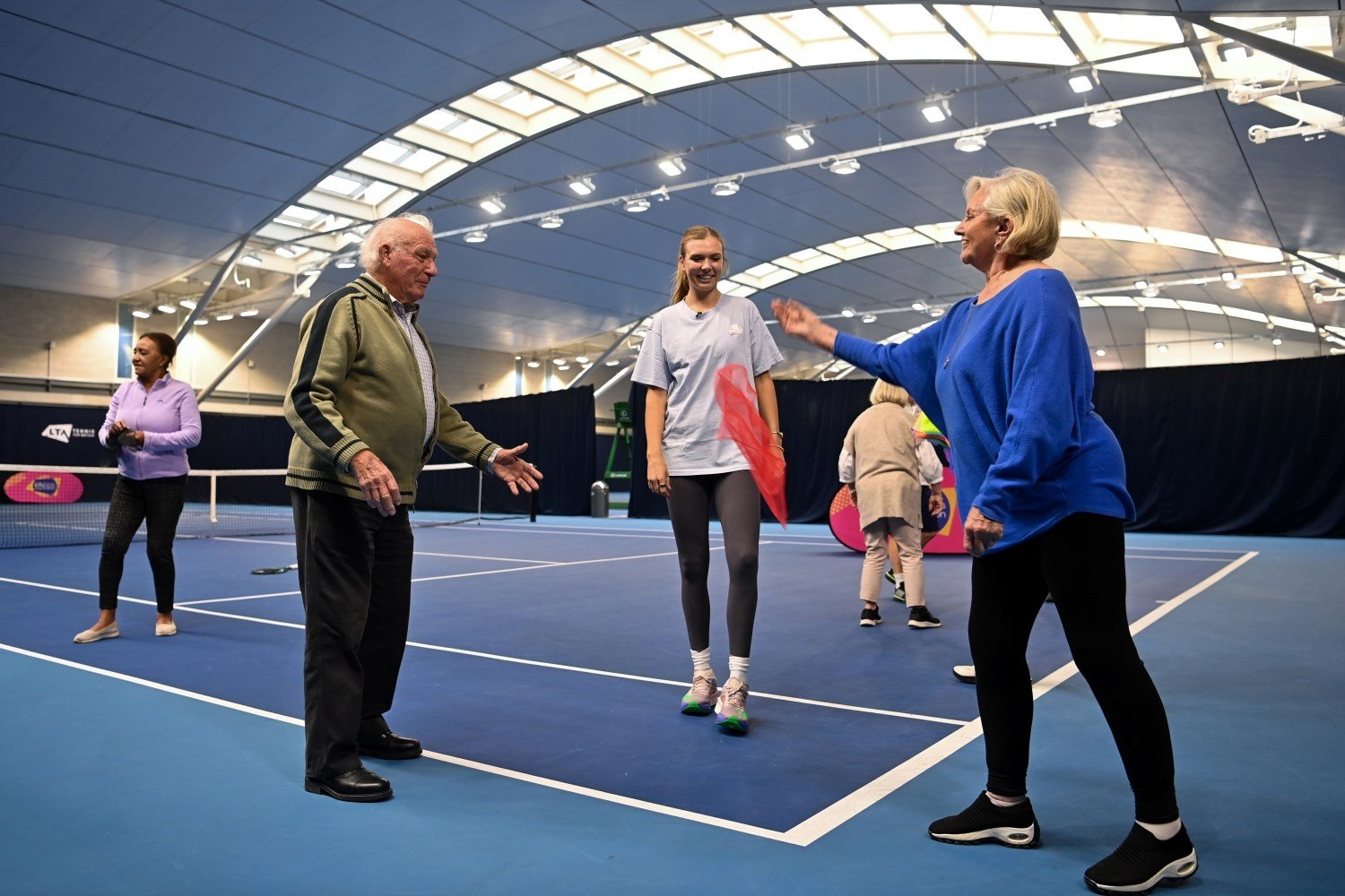 A group of three people - an old man, and old woman, and a young woman between the other two - stand on a tennis court, throwing a red scarf between them. 