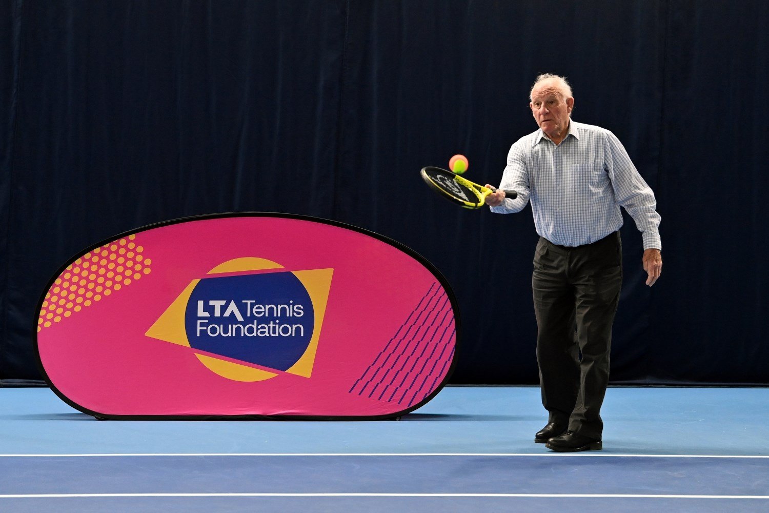 An old manhit a red and yellow tennis ball with a tennis racket. Next to him is a pink sign with the words, 'LTA Tennis Foundation' written on it