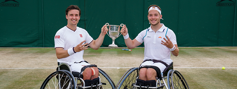 Alfie Hewett and Gordon Reid holding the Wimbledon trophy together on a tennis court