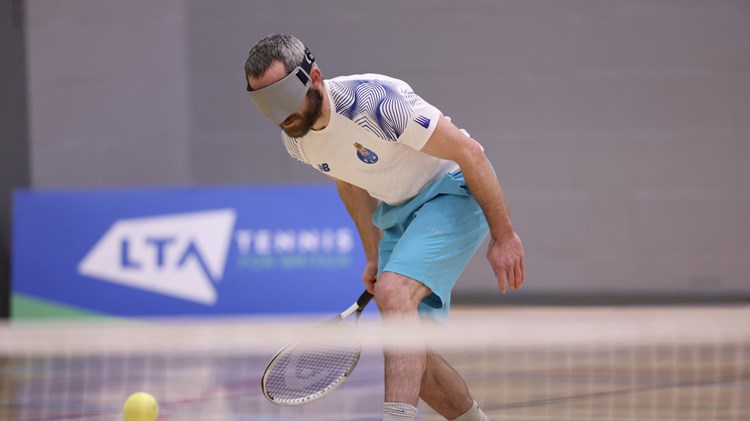 A man wears a grey blindfold and sport kit, as he tries to hit a tennis ball with a tennis racquet. A tennis net is blurred in the foreground, and a blue sign is visible in the background wioth the letters 'LTA' on it