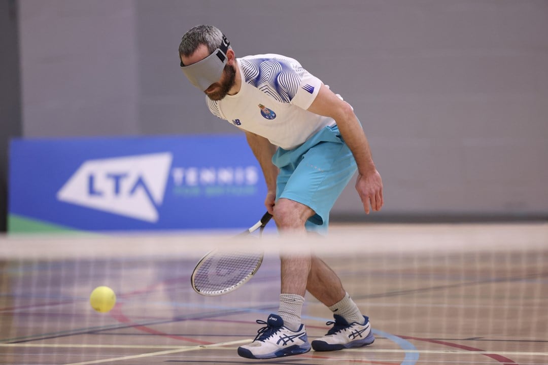 A man wears a grey blindfold and sport kit, as he tries to hit a tennis ball with a tennis racquet. A tennis net is blurred in the foreground, and a blue sign is visible in the background wioth the letters 'LTA' on it
