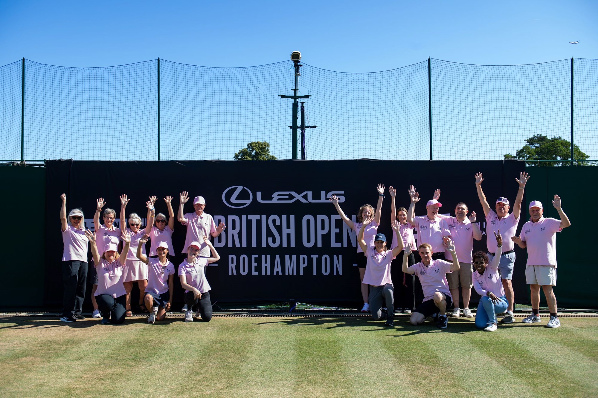 The group of Lexus British Open volunteers posing in two groups with their hands in the air next to the event sign
