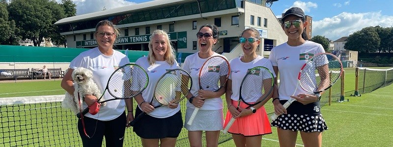 A group of five women smiling together on an outdoor tennis court with their tennis rackets and a dog