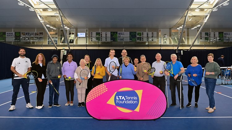 A group of elderley people, with a young woman in the middle of the grop and a younger man at the far left of the group, stand on a tennis court holding tennis rackets, with a pink sign in front of them with the words, 'LTA Tennis Foundation' on it.