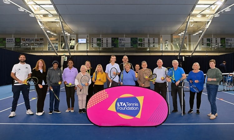 A group of elderley people, with a young woman in the middle of the grop and a younger man at the far left of the group, stand on a tennis court holding tennis rackets, with a pink sign in front of them with the words, 'LTA Tennis Foundation' on it.