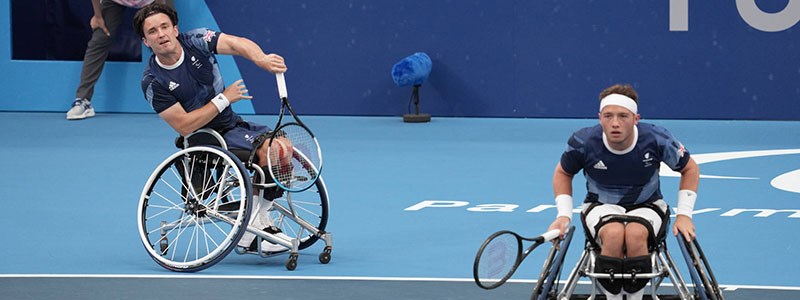 Two wheelchair tennis players mid game returning a shot