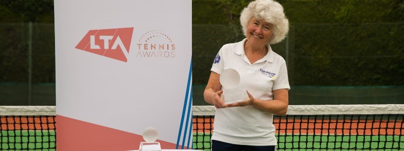 Sarah Strawbridge holding an award on an outdoor tennis court in front of an LTA tennis award board