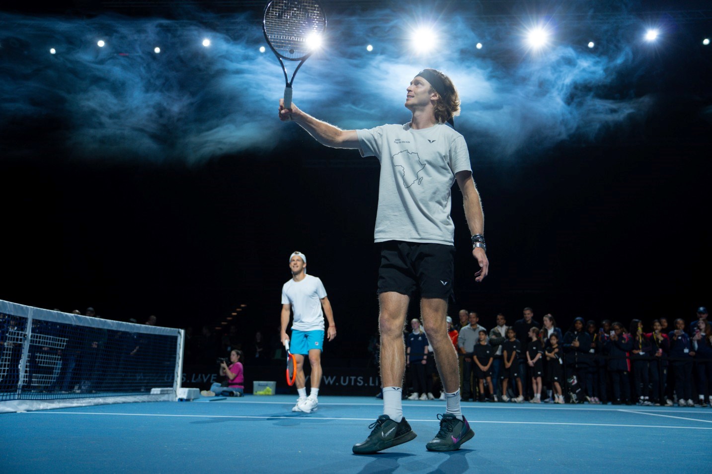 Tennis player Andrey Rublev stood on court at the Ultimate Tennis Showdown, holding his racket up to the sky while ball kids look on at him