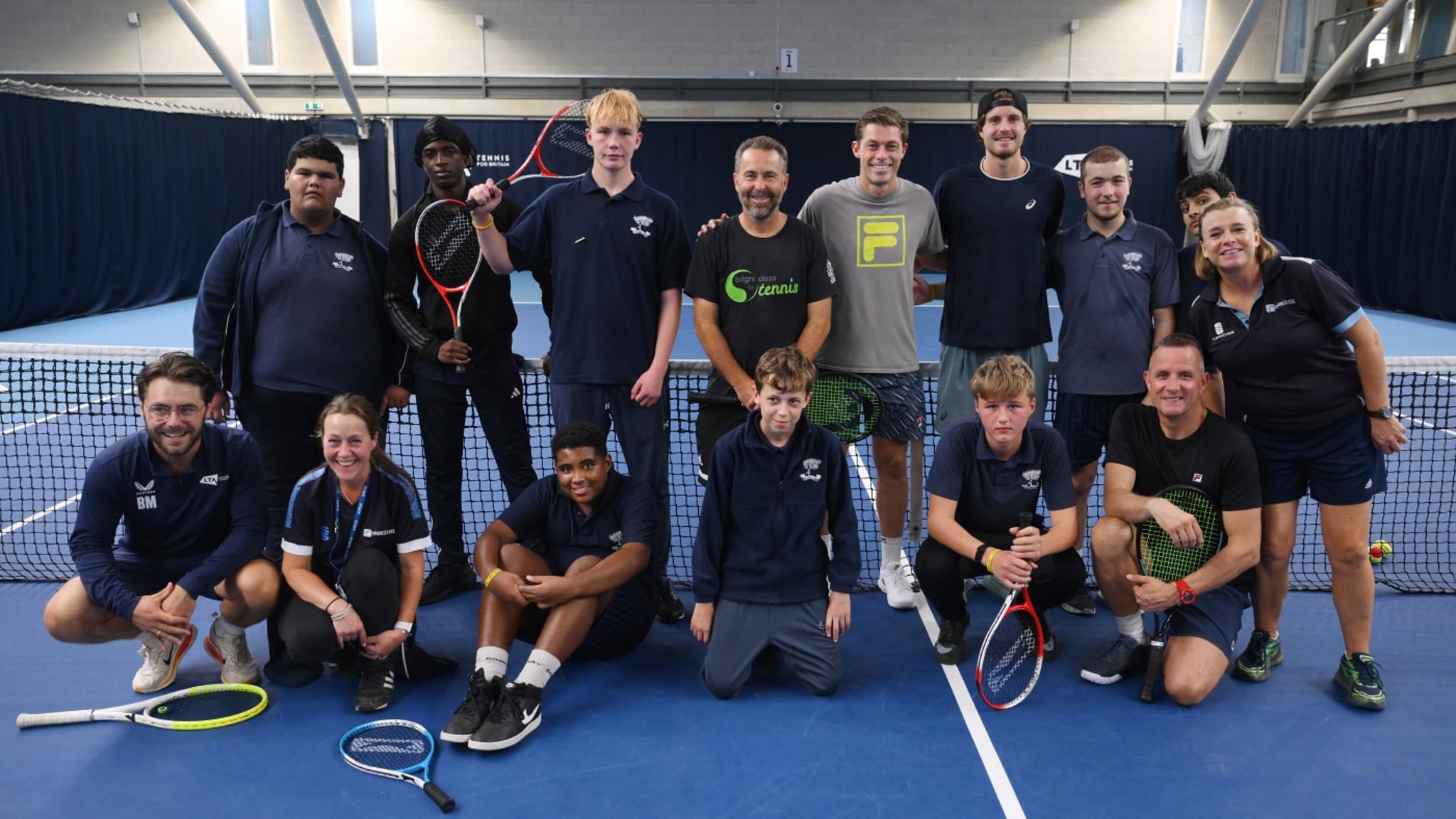 A group of 9 young people with two coaches stood in front of the net on a tennis court with professional tennis players Neil Skupski and Tobey Samuel