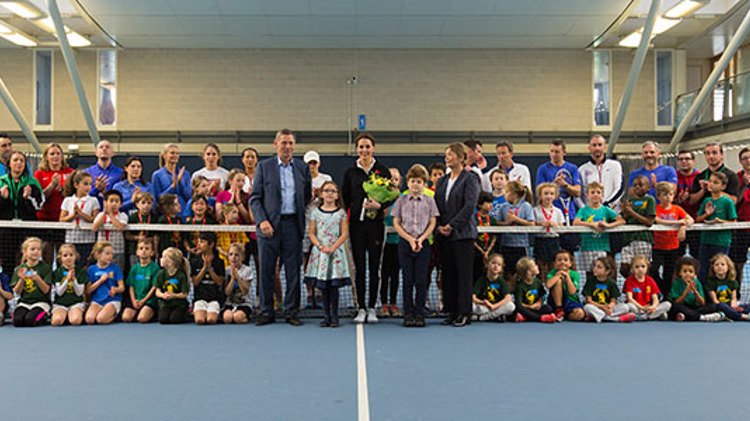 Duchess of Cambridge standing in the middle of a group of kids and coaches on a tennis court by the net
