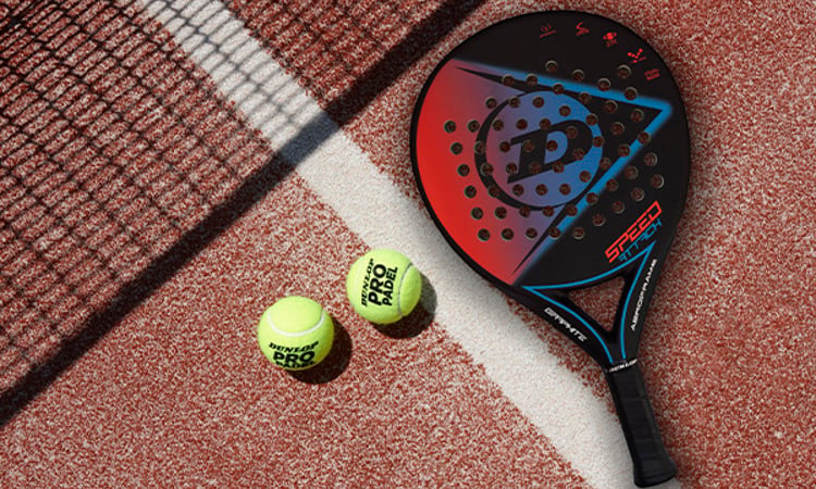 Two padel balls and a Dunlop padel bat on a clay-coloured padel court
