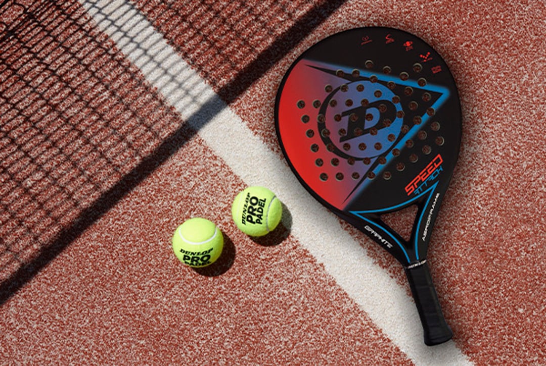 Two padel balls and a Dunlop padel bat on a clay-coloured padel court