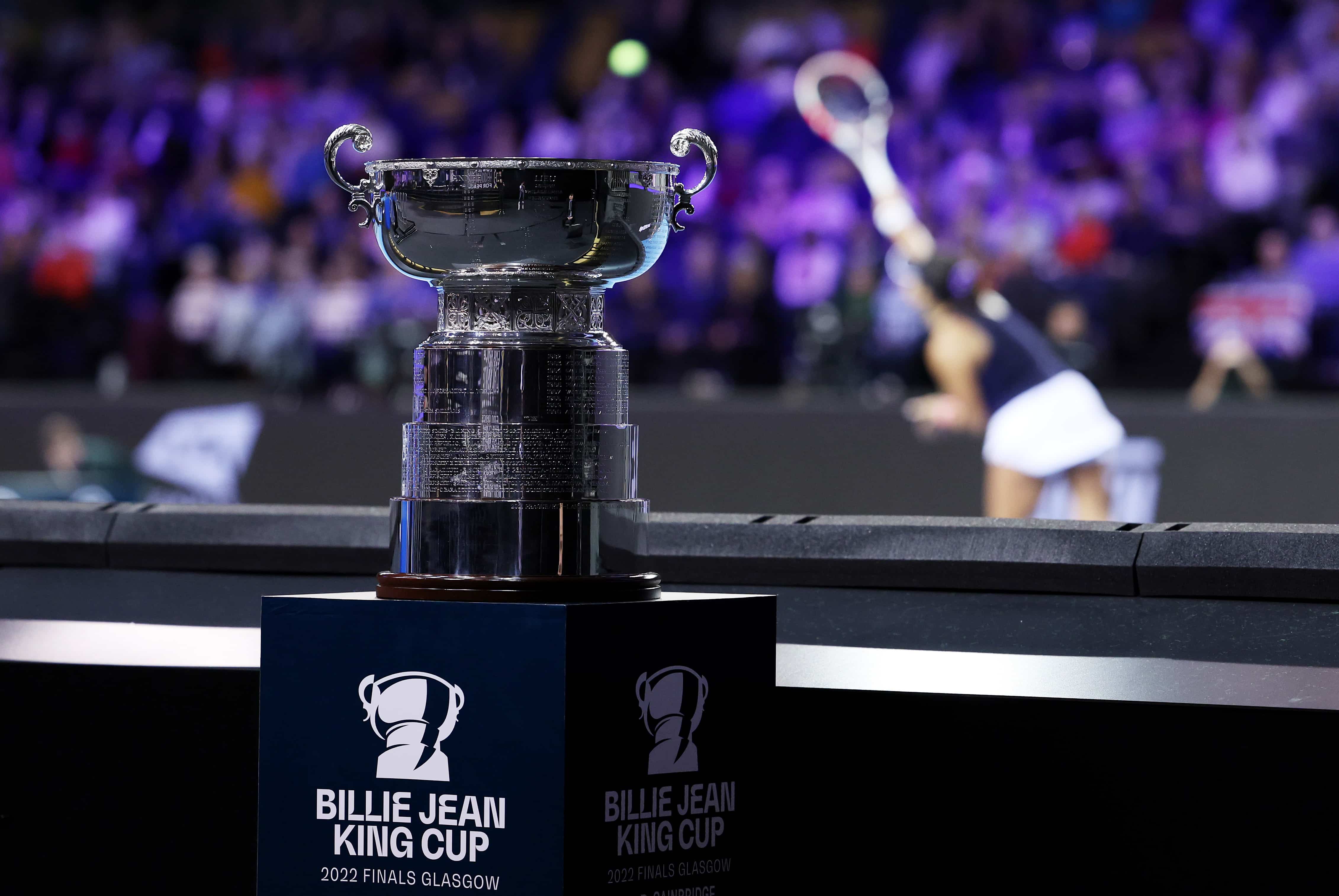 The Billie Jean King Cup 2022 trophy on its stand in front of a tennis player serving