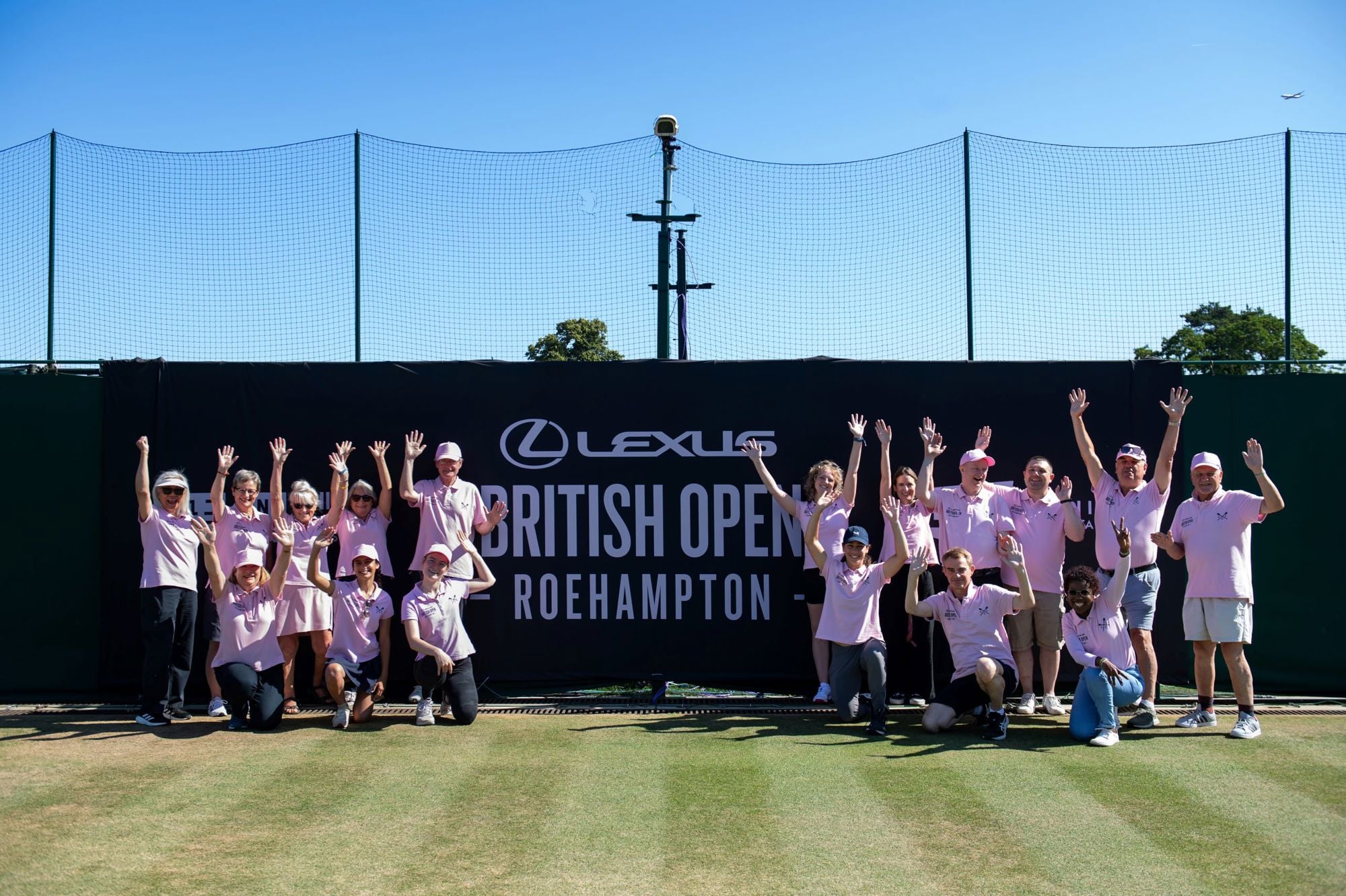 The group of Lexus British Open volunteers posing in two groups with their hands in the air next to the event sign