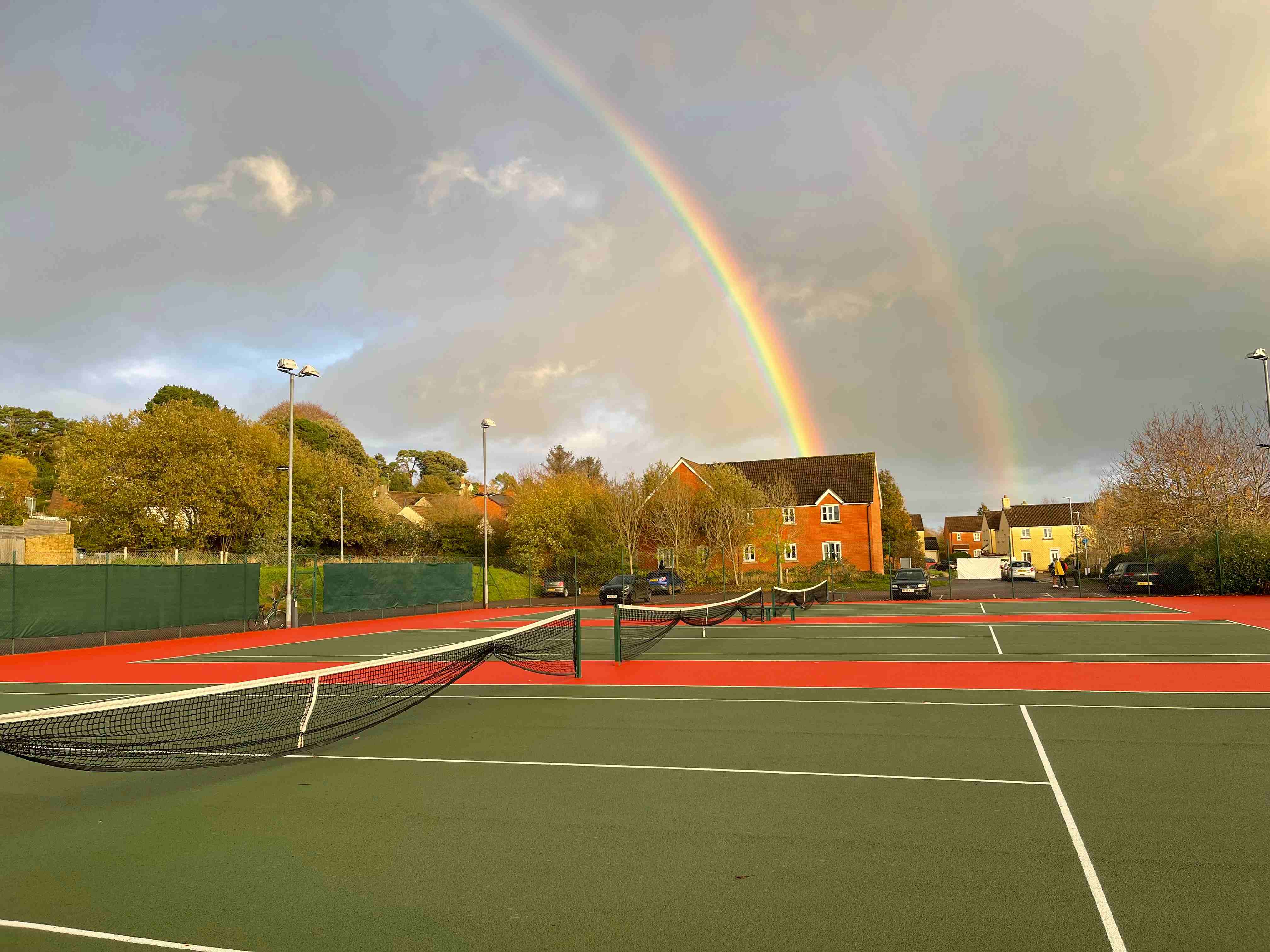 A picture of three tennis courts with houses behind them and a rainbow in the sky