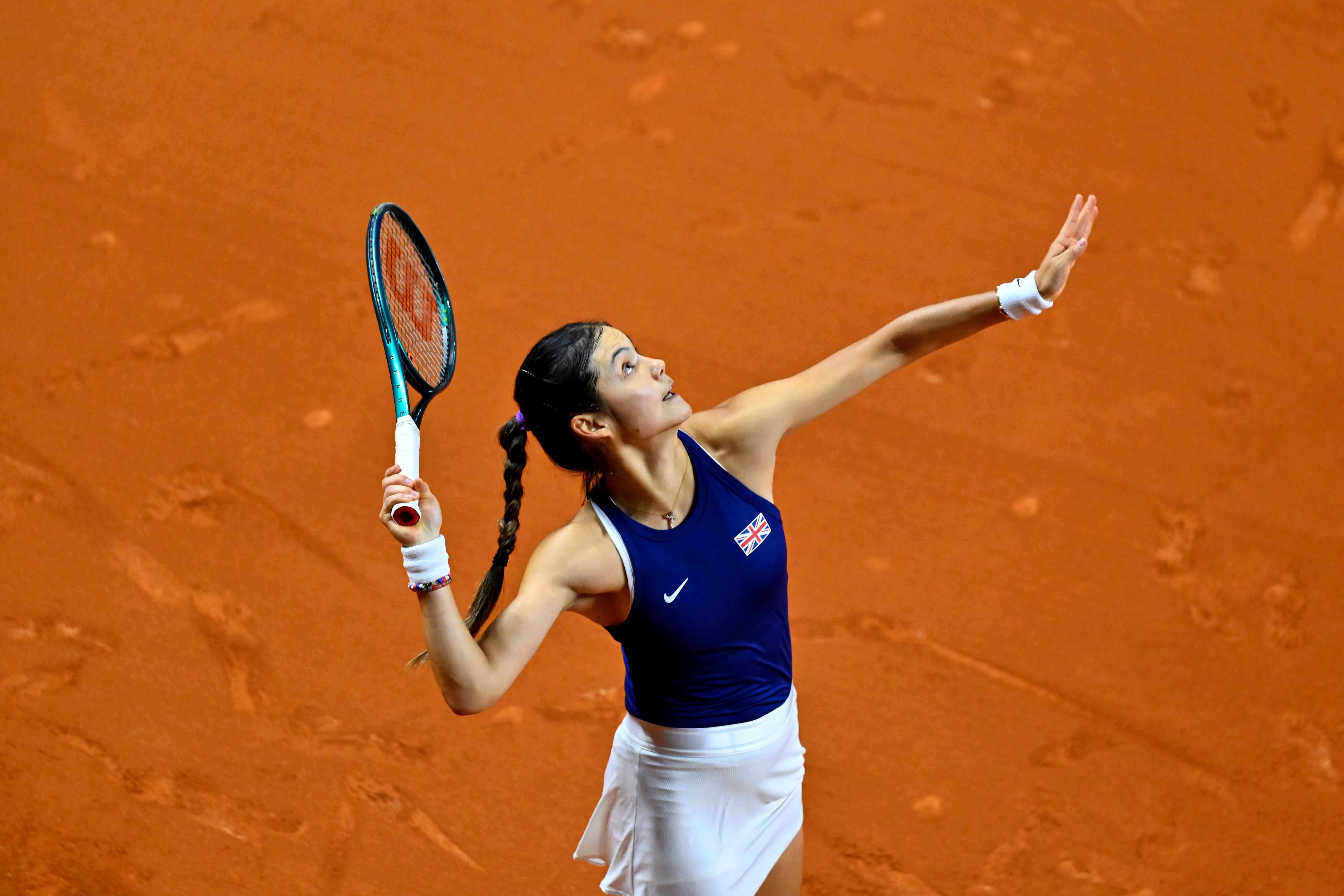 Emma Raducanu tossing a tennis ball in the air while preparing to serve on an orange clay court 