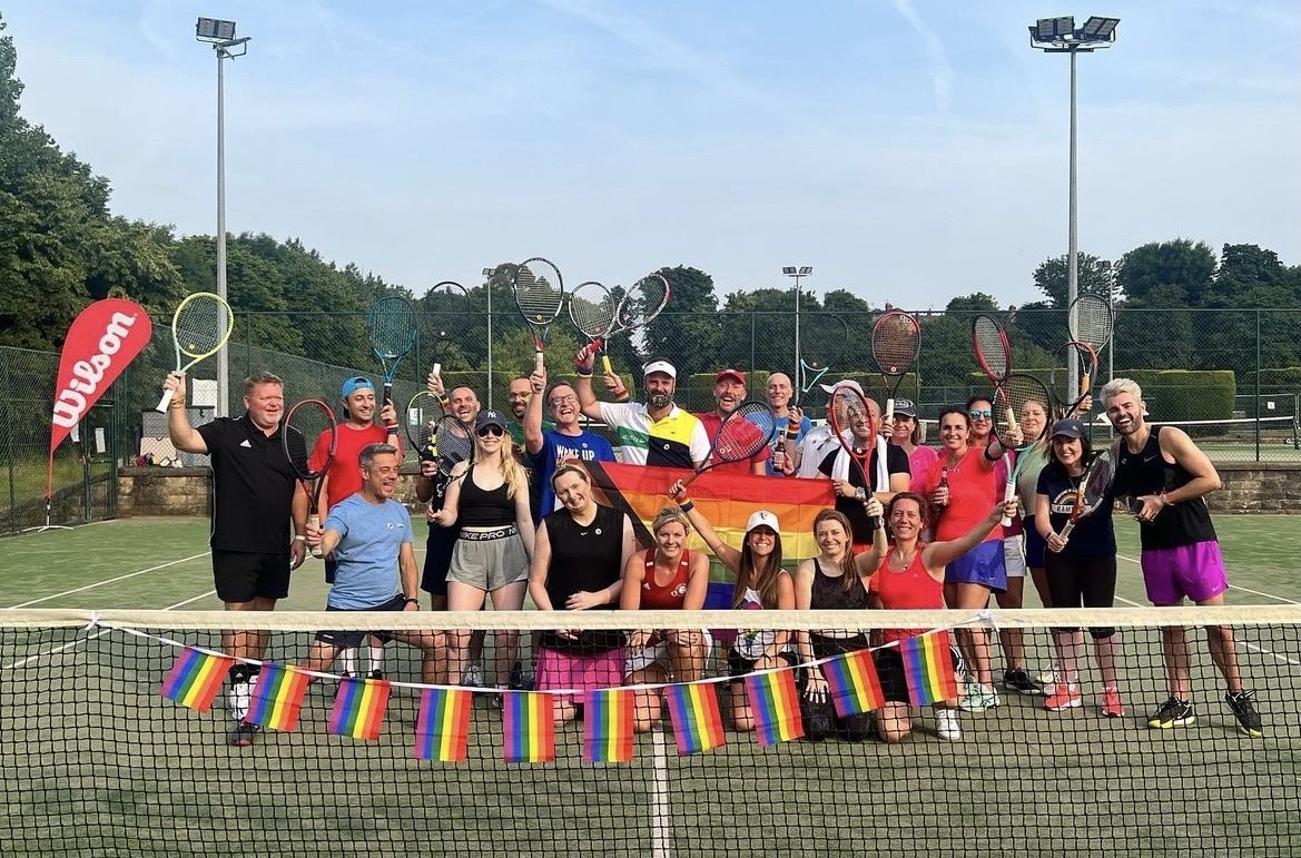 A large group of people standing on a tennis court, posing with tennis rackets dressed in brightly coloured clothing, behind a net draped with rainbow flags