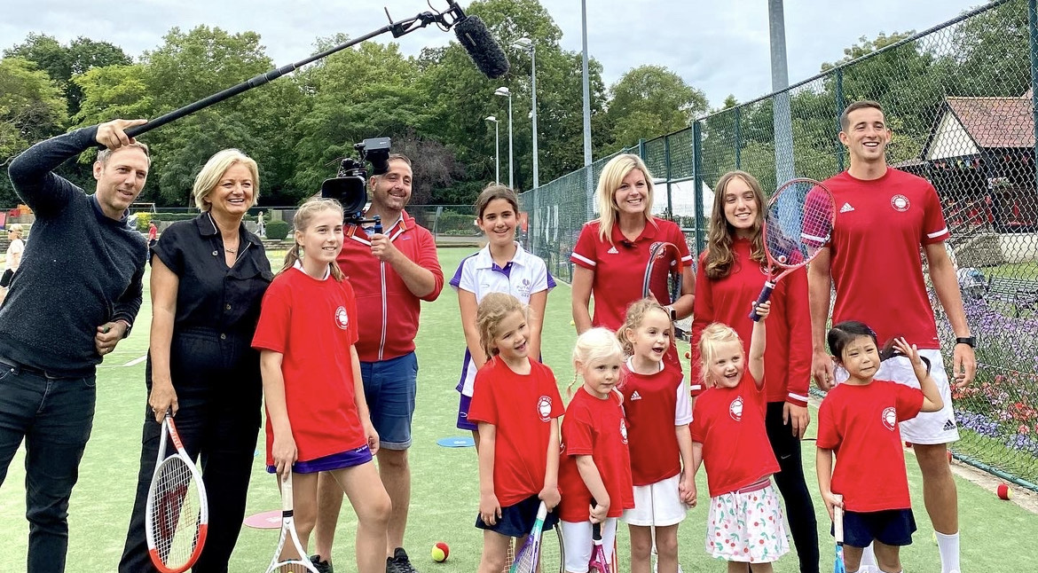 A group of children and adults posing with tennis rackets, with a man holding a large microphone and a man holding a tv camera stood next to them