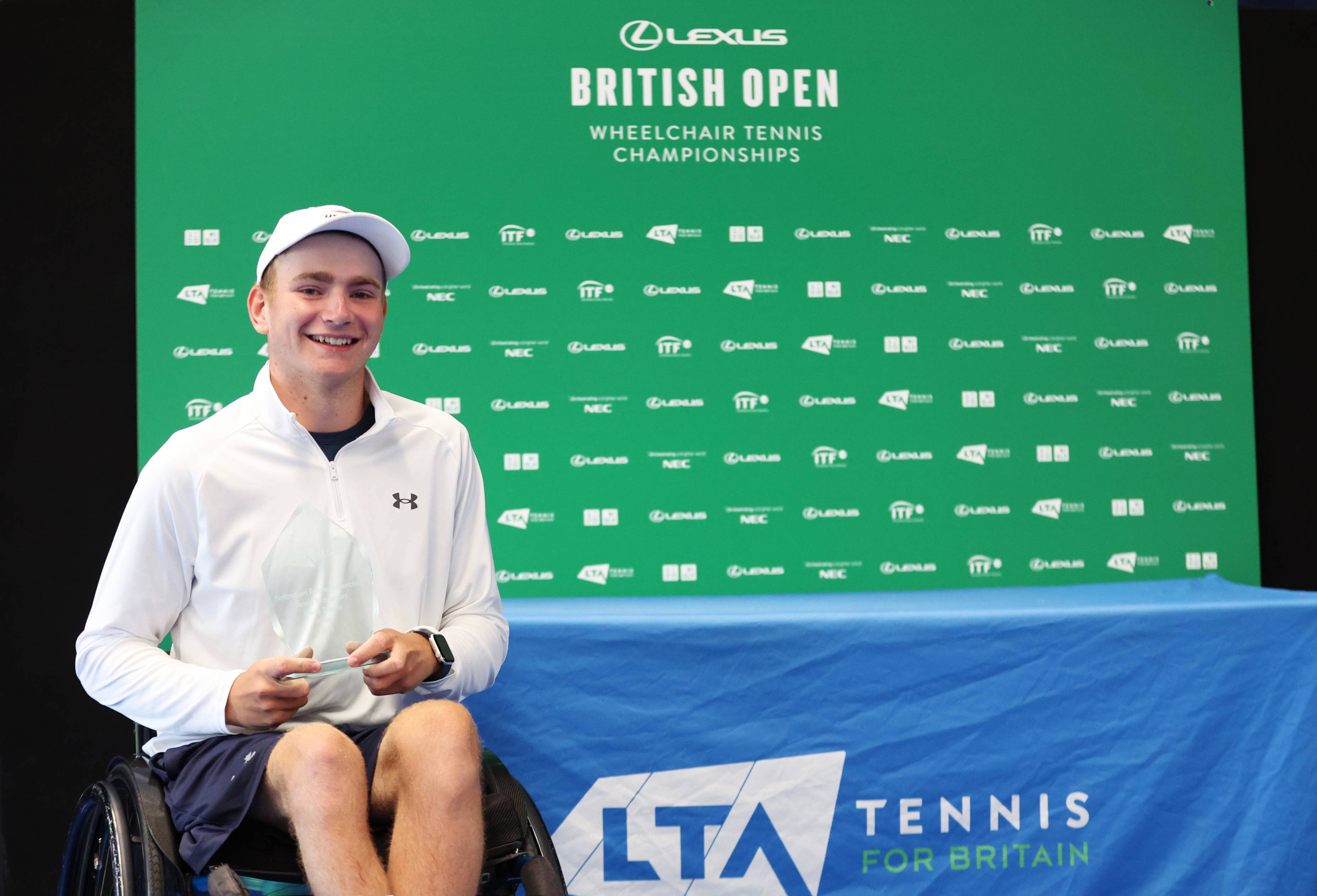 British wheelchair tennis player Andrew Penney smiling while sat in his wheelchair in front of a 'Lexus British Open Wheelchair Tennis Championships' branding board