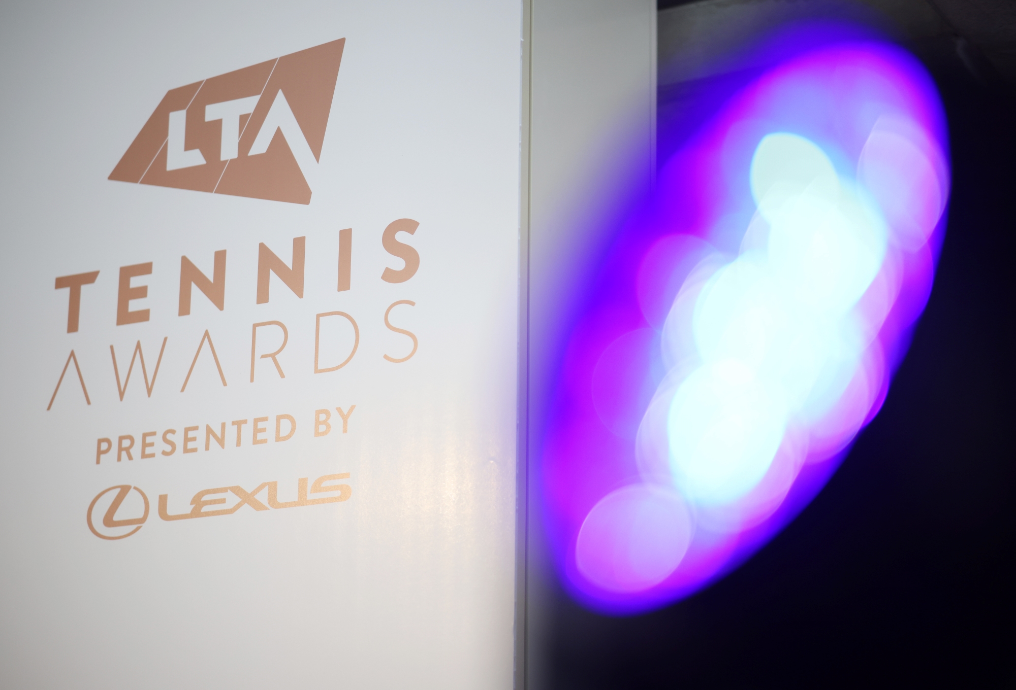 The text, 'LTA Tennis Awards presented by Lexus', in gold font on a white background, with a purple light out of focus to the right hand side