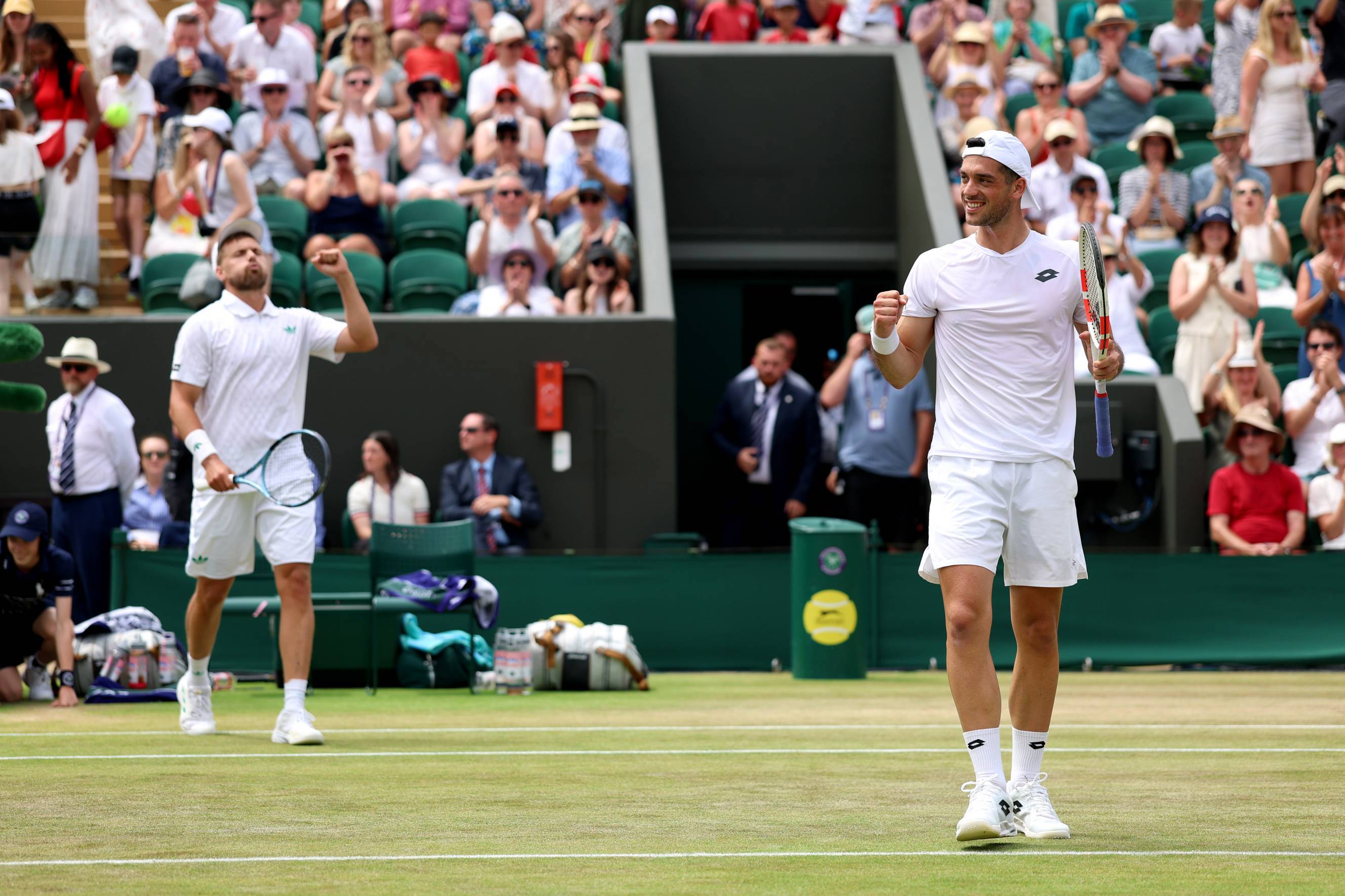 Julian Cash pumping his fist and racket in celebration while Lloyd Glasspool is behind him raising his fist in celebration in front of the Wimbledon crowd