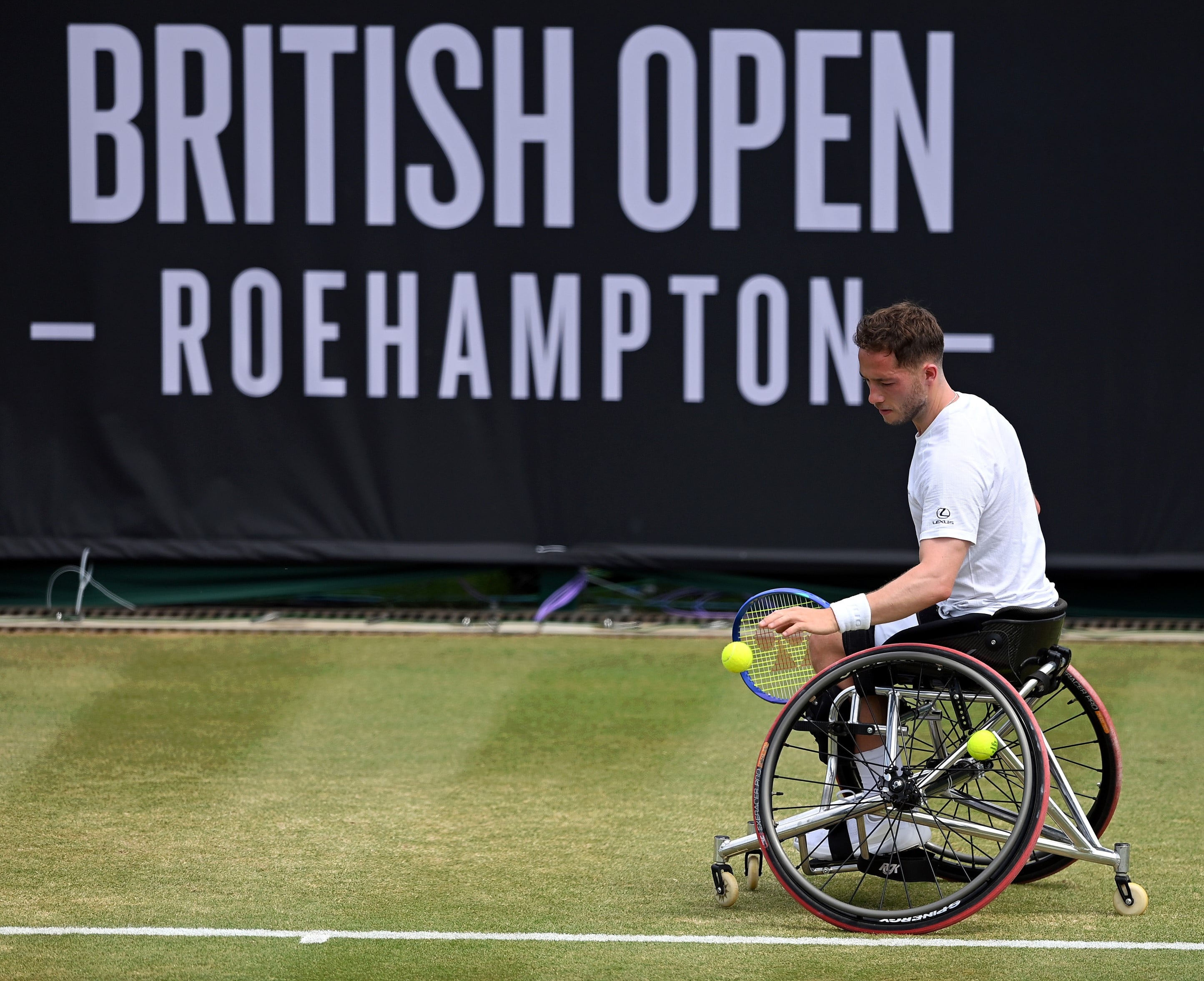Alfie Hewett bouncing the ball before a serve on grass in front of the Lexus British Open sign
