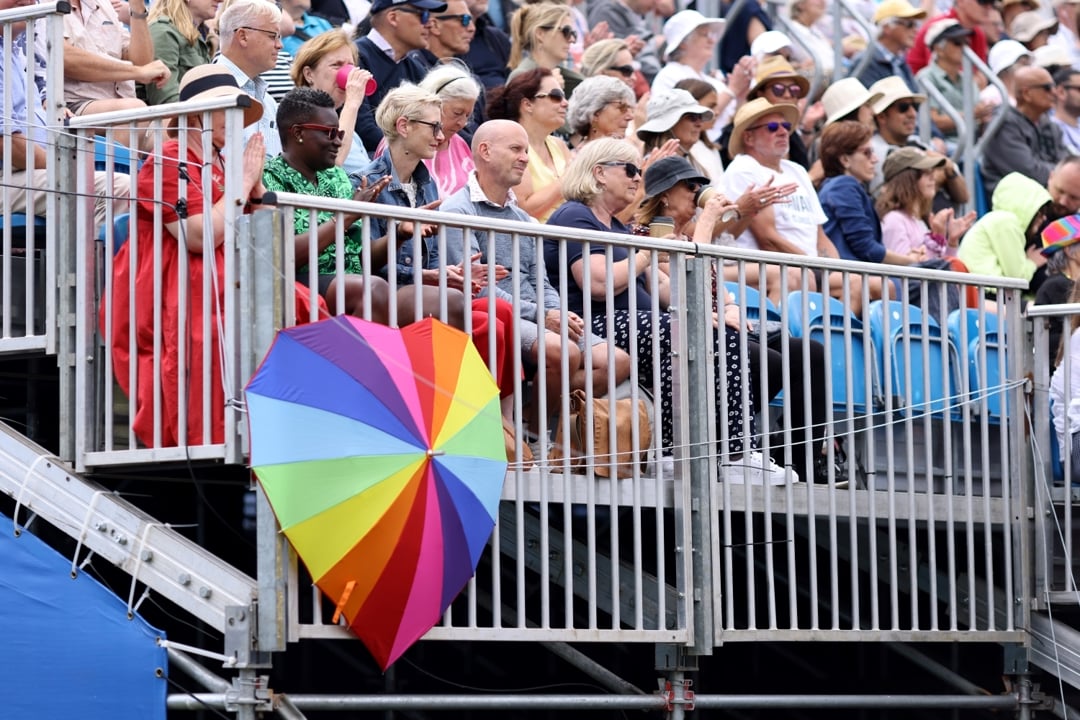 A multi-coloured umbrella is open, in the shape of a heart, resting in front of railing, with spectators sat behind