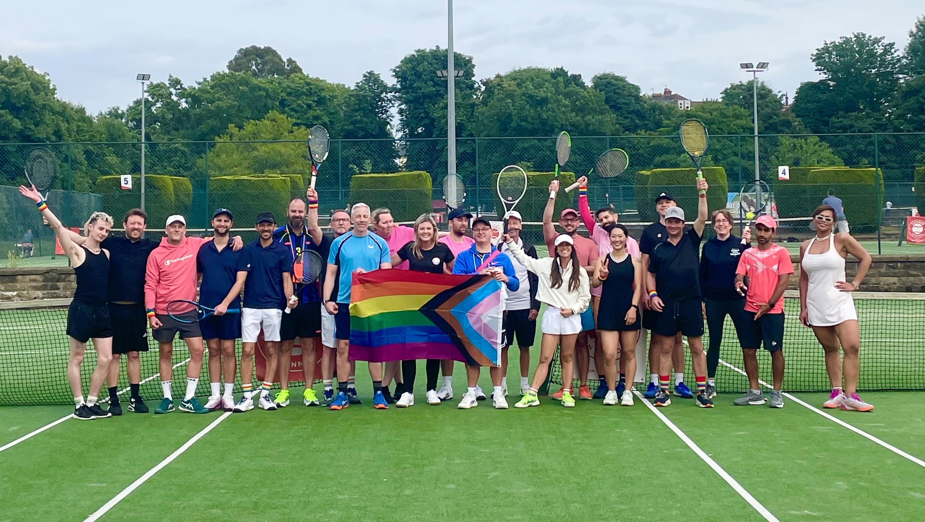 A group of people stand on a tennis court, holding tennis rackets in the air . In the middle of the group three people between them hold a Pride flag