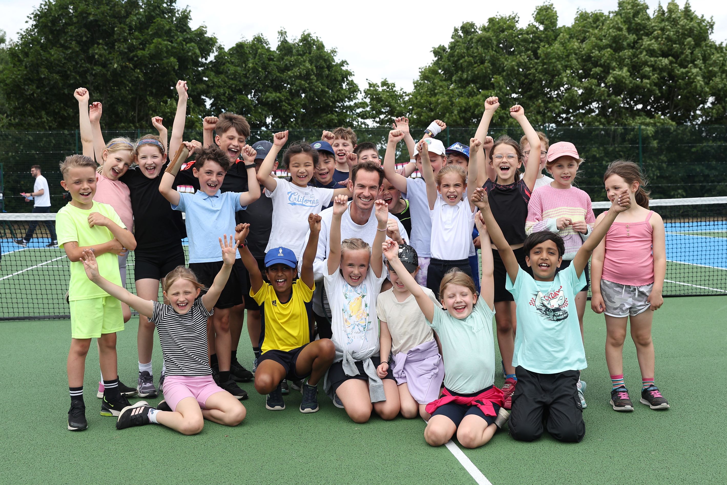 Andy Murray smiling in the middle of a group of young kids cheering with their hands in the air on a park court
