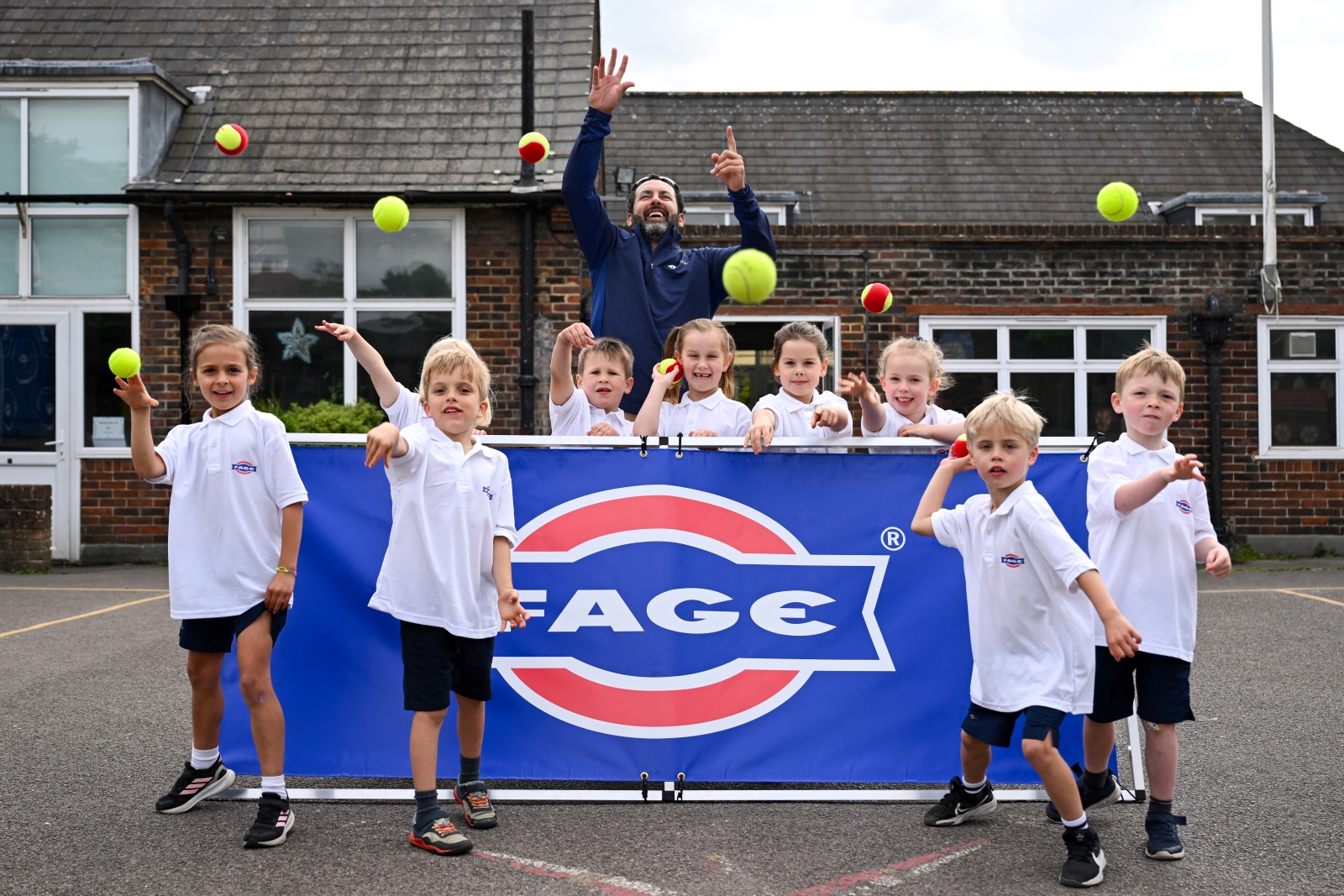A group of children, stood in front and behind of a  blue banner with the word 'FAGE' on it, throw tennis balls towards the camera. There is an adult man stood with the group as well.