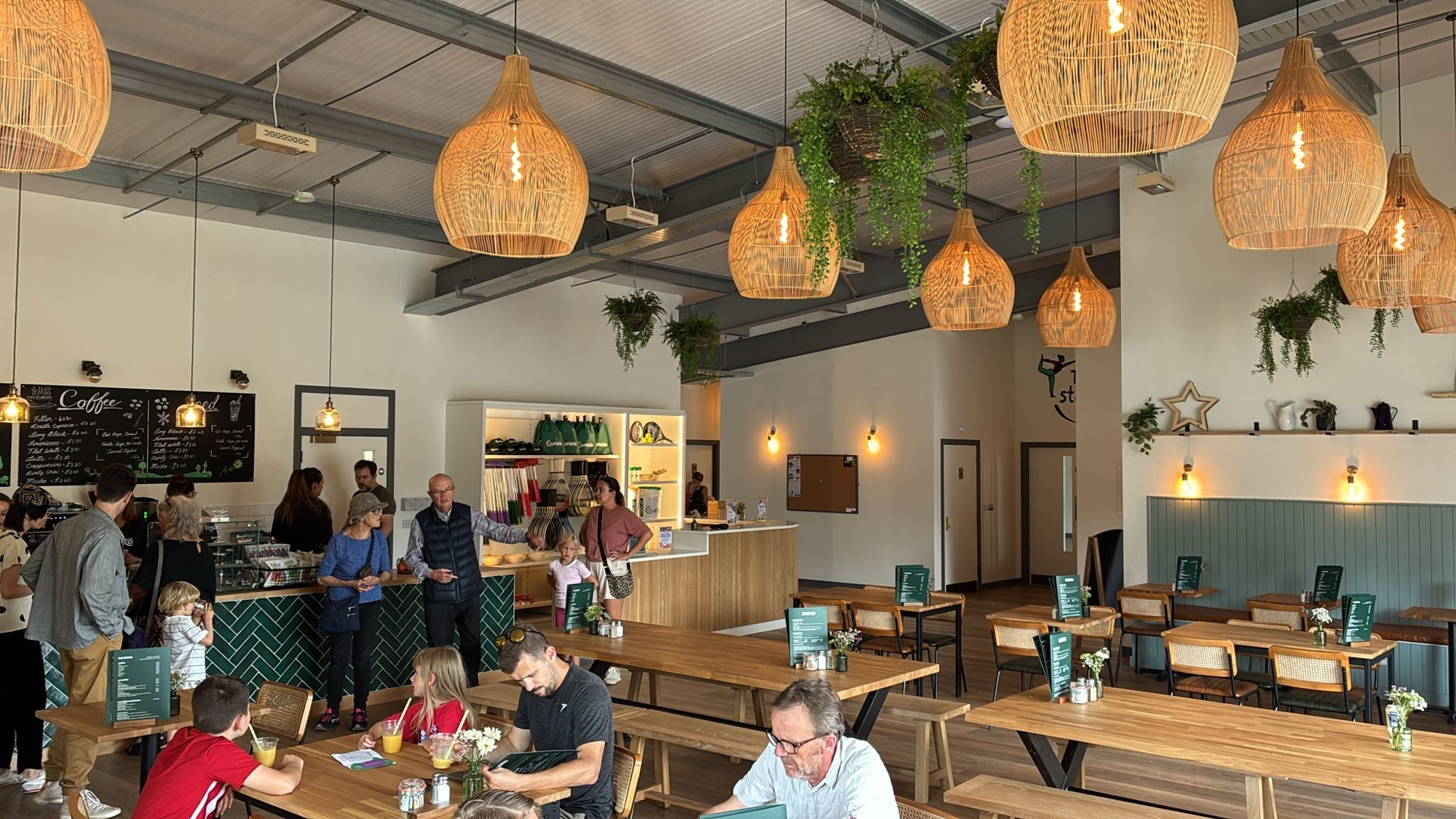 A large open plan room filled with wooden tables and benches. Hangin from the ceiling are lights with large shades on them. At the back of the room, people queue at a counter stacked with food offerings, and on the back wall a blackboard with 'Coffee' written in large letters displays menu options
