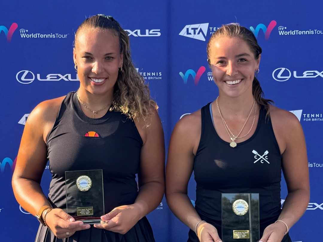 Two women, both wearing black vest tops, stand smiling and holding glass trophies. Behind them is a blue backdrop featuring sponsor logos