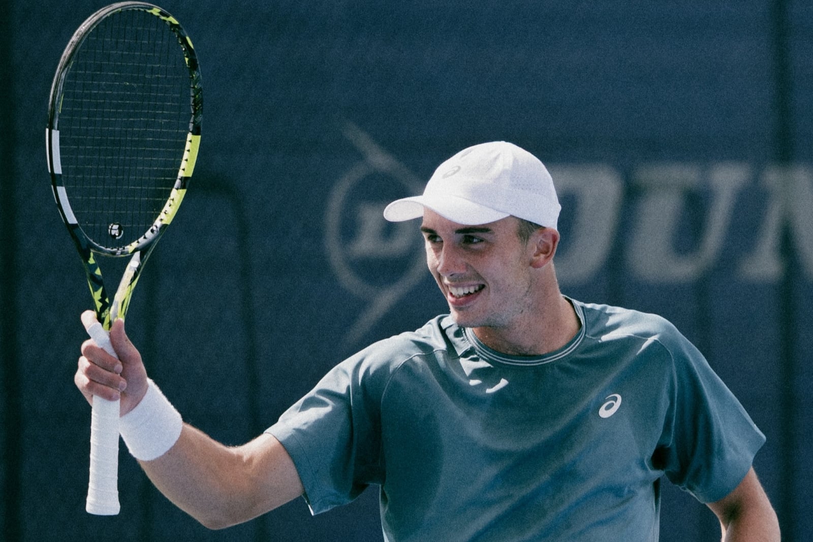 A man wearing a grey t-shirt, a white sweatband on his right wrist and a white cap, smiles as he holds up a tennis racket