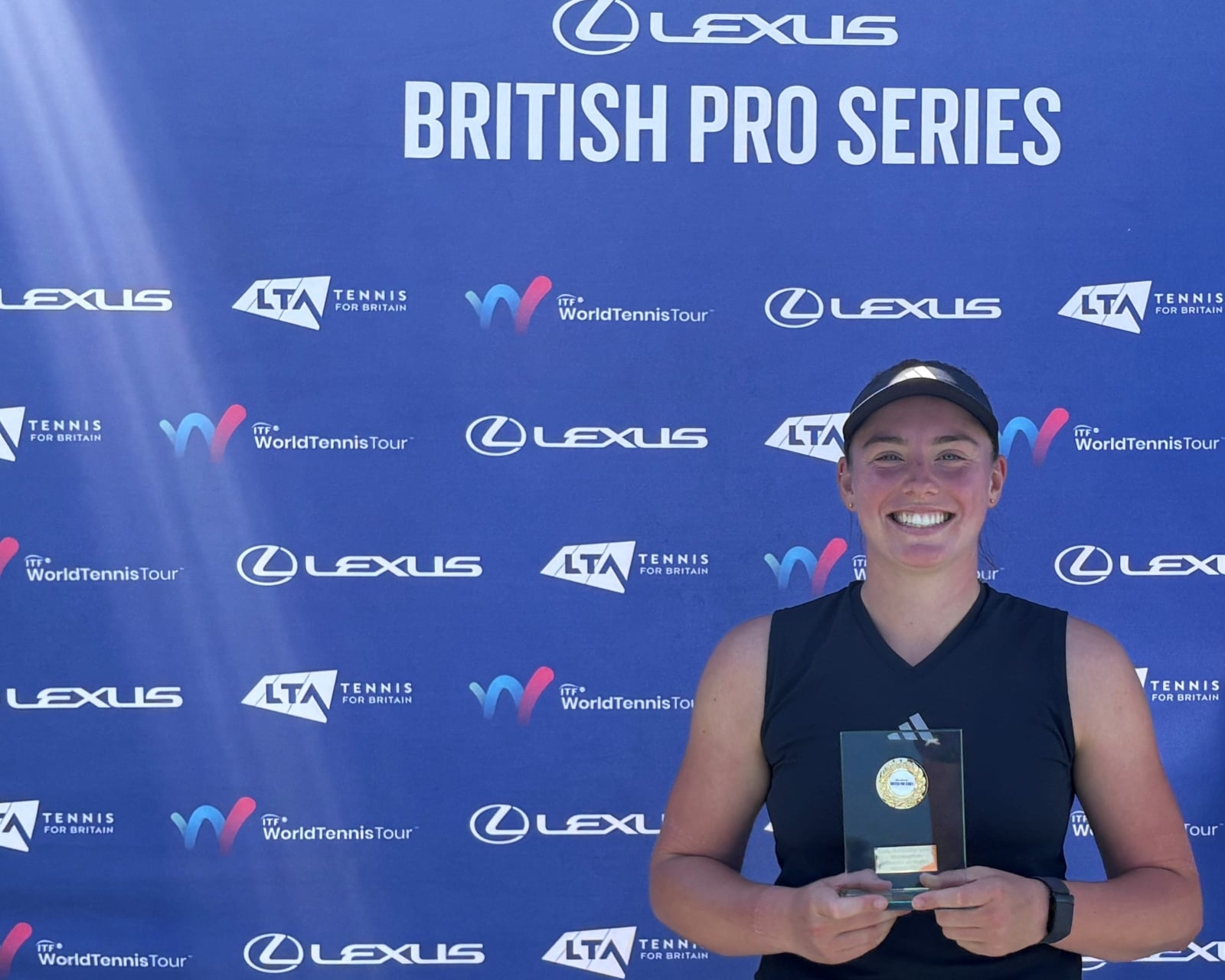 A woman wearing a black vest top holds a glass trophy, smiling. Behind her is a blue sponsor board with the words, 'Lexus British Pro Series' visible, along with a variety of sponsor logos