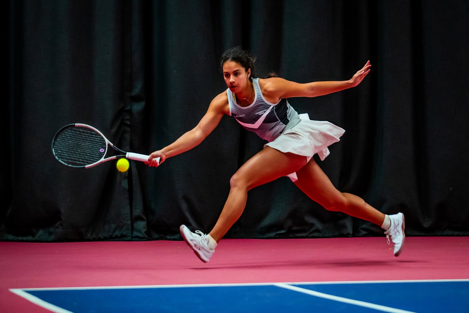 A woman runs to hit a tennis ball. She is wearing a grey vest top, a white skort and white trainers. The edge of a blue tennis court surface is visible, while she runs on a pink coloured floor outside of the court lines