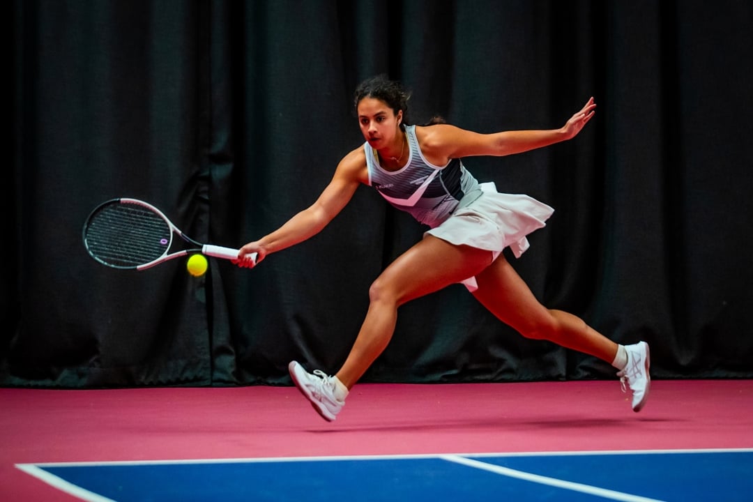 A woman runs to hit a tennis ball. She is wearing a grey vest top, a white skort and white trainers. The edge of a blue tennis court surface is visible, while she runs on a pink coloured floor outside of the court lines