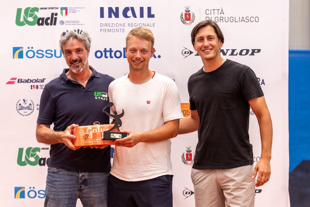 Three men stand side by side, with the man in the middle, wearing a white-t-shirt and black shorts, holding a black trophy